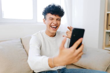 Kanepede oturan neşeli bir genç, akıllı telefonuyla mutlu bir selfie çekiyor. Parlak, doğal ışık, iç mekan ortamının neşeli atmosferini arttırıyor.