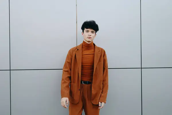 Stock image A young man stands confidently in a brown suit with a turtleneck, set against a neutral gray backdrop The scene emphasizes contemporary fashion and individuality