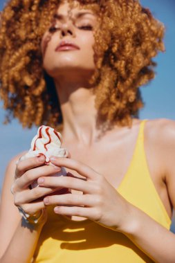 Kıvırcık saçlı kendine güvenen bir kadın dondurmadan hoşlanır. Güneşli bir günde parlak sarı bir mayo giyer.