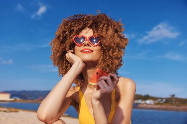 Sarı mayo giymiş neşeli genç bir kadın sahilde bir dilim karpuz, arka planda parlak mavi gökyüzü ile poz veriyor.