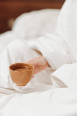 Bir fincan çay ve battaniyeyle yatakta oturan kişi. Sıcak bir içecekle yatakta sıcak ve samimi kal. Rahatlamak ve rahatlamak için mükemmel bir görüntü..