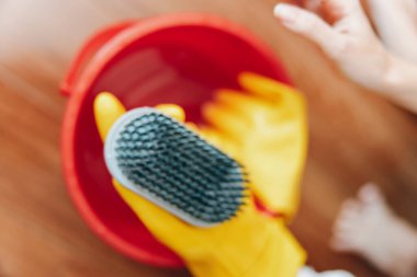 Person wearing yellow rubber gloves holds a brush and a red bowl on a wooden floor. clipart