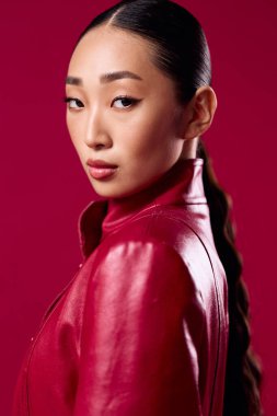 Kırmızı deri ceketli Asyalı kadın ve pembe arka planda at kuyruklu örgü. Photoshoot 'a poz veren esmer model. Genç kadınlar için modaya uygun ve şık görünüm.