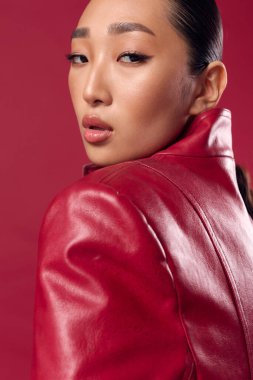 Kırmızı deri ceketli Asyalı esmer kadın kırmızı arka planda bir fotoğraf için poz veriyor..
