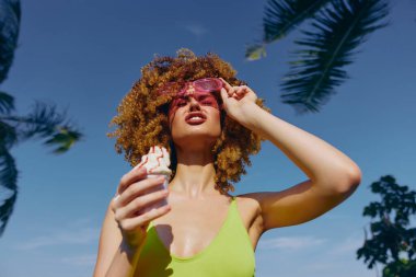 Canlı bir yaz sahnesi. Kıvırcık saçlı, dondurmanın tadını çıkaran, şık pembe güneş gözlüğü takan ve parlak bir mayo giyen genç bir kadın.