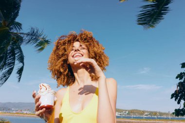 Kıvırcık saçlı neşeli bir kadın dondurma bardağını tutuyor, açık mavi gökyüzüne gülüyor ve tropikal arka planda