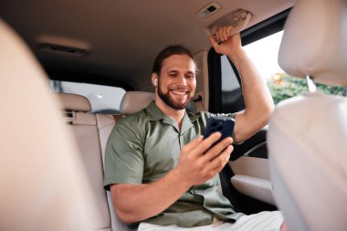 Yeşil tişörtlü neşeli bir adam, kulaklıkları takılı, akıllı telefonunu bir arabanın arka koltuğunda kullanıyor, rahatlama ve zevk somutlaştırıyor.