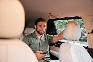 Kulaklıklı rahat bir adam arabanın arka koltuğunda oturmuş, yemyeşil bir arka planda akıllı telefonunun keyfini çıkarıyor.