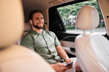 Yeşil tişörtlü ve şortlu rahat bir adam arabanın arka koltuğunda oturuyor. Telefonunu kablosuz kulaklıklarıyla gülümsüyor.