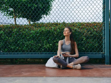Spor kıyafetleri içinde dingin genç bir kadın sarayda bacak bacak üstüne atarak oturuyor, çevresinden zevk alırken gülümsüyor, arka planda yemyeşil bir ortam var.
