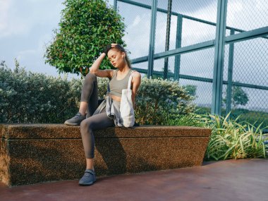 Sportif bir kadın model, sakin bir açık hava ortamında, yemyeşil bitkiler ve güzel bir gökyüzü içeren bir bankta gelişigüzel poz veriyor.