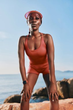 Kırmızı spor kıyafetli fit bir kadın arkasında açık mavi gökyüzü ve sakin bir denizle kayalık bir arazide duruyor.