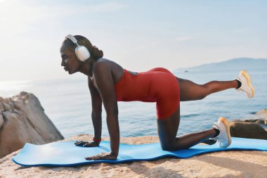 Kırmızı spor kıyafetli bir kadın, sakin denize bakan bir mindere yoga pozu veriyor, kulaklık takıyor ve spor salonuna odaklanıyor.