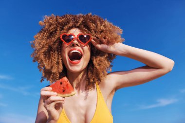 Neşeli bir kadın, açık mavi bir gökyüzünün altında, sarı bir mayo giymiş bir karpuz dilimini tutarken geniş bir gülümseme sergiliyor.