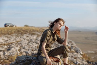 Kadın, arka planda araba, seyahat ve macera konseptiyle bir kayanın tepesine kafa yoruyor.