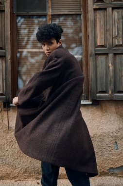 Bir binanın önünde duran kahverengi ceketli şık bir gencin moda fotoğrafı. Başında bir şapka ve boynunda bir eşarp var..