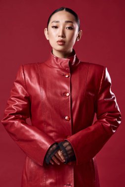 Kırmızı deri ceketli esmer, Asyalı kadın elleri kalçasında kırmızı arka planda poz veriyor. moda konsepti.