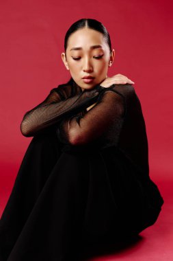 Siyah bluzlu ve pantolonlu Asyalı bir kadın kırmızı arka planda oturuyor elleri dizlerinde ve kafası dizlerinin üzerinde dinleniyor. moda konsepti.