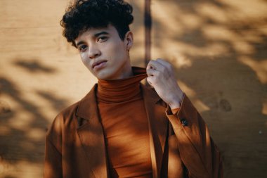 Kahverengi balıkçı yaka ve ceket giyen kıvırcık saçlı genç bir adam, sıcak bir ahşap zemine nazikçe poz veriyor.