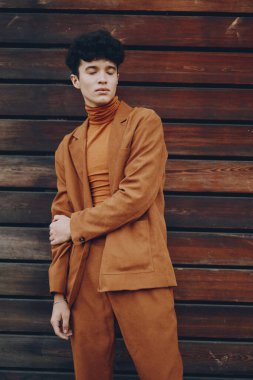 Modaya uygun bir erkek model, doğal ahşap zemin ile modern eğilimleri gözler önüne seren tek renkli kahverengi bir kıyafet içinde zarif bir poz verir.