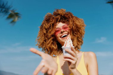 Kıvırcık saçlı, dondurma tutan, güneş gözlüğü takan, parlak bir arka planda gülümseyen mutlu bir genç kadın.