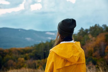 Şık sarı yağmurluklu bir kadın sonbahar gezisi boyunca nefes kesen dağ manzarasına hayran.