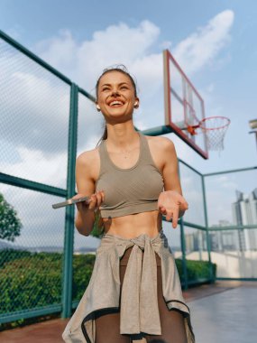 Akıllı telefonlu mutlu atletik kadın, canlı bir geçmişi olan basketbol sahasında eğleniyor.