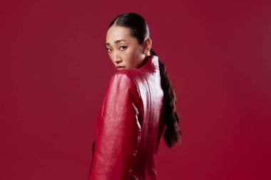 Kırmızı deri ceketli Asyalı manken elleri kalçasında kırmızı arka planda poz veriyor. şık ve kendine güvenen güzellik.