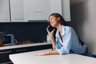 Mutfak tezgahında oturan mutlu kadın cep telefonuyla gülümsüyor.