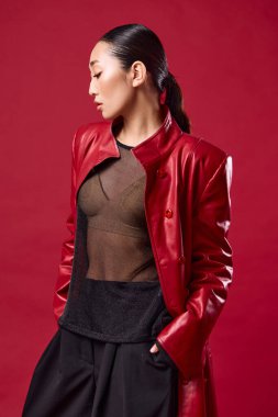 Kırmızı deri ceketli, siyah pantolonlu, elleri kalçasında kırmızı arka planda poz veren Asyalı esmer kadın. Saçını at kuyruğuyla arkaya çekti..