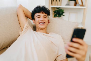 Gülümseyen genç adam rahat bir şekilde koltukta oturuyor, akıllı telefonunun keyfini çıkarırken, şık bir oturma odasında minimalist bir dekor içinde keyif ve rahatlık hissi ile dinleniyor.