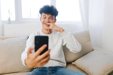 Mutlu genç adam gülümsüyor ve akıllı telefonunda selfie için poz veriyor. Açık renkli bir kanepede rahat bir şekilde oturuyor.