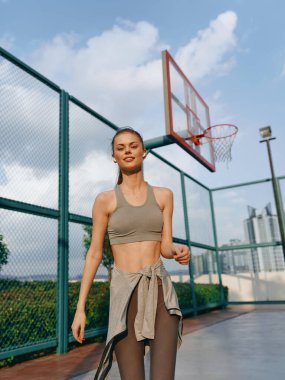 Kendine güvenen genç bir kadın yeşil ağaçlarla çevrili parlak ışıklı bir basketbol sahasında yürüyor.
