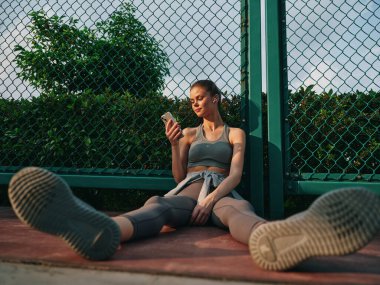 Bir fitness meraklısı telefonunu kontrol ederken rahatlıyor, refah ve teknolojiye odaklanmış sıradan bir açık hava havası sergiliyor.