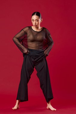 Siyah pantolonlu Asyalı bir manken ve parlak kırmızı arka planda elleri kalçalarının üzerinde durarak güvenle poz veriyor. şık ve zarif..