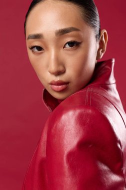 Kırmızı arka planda poz veren kırmızı deri ceketli Asyalı kadın. Modaya uygun esmer model kameraya bakıyor. Moda ve şık fotoğraf çekimi konsepti.