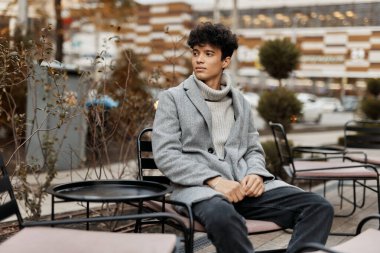 Gri montlu, şık bir genç adam, şehrin arka planında, ağaçlı bir sandalyede oturuyor. Modern ortamdaki şık şehirli adam..