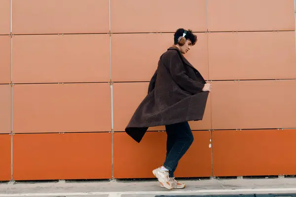 stock image Asian woman in brown coat and black pants walking in front of orange brick wall. fashionable urban street style.