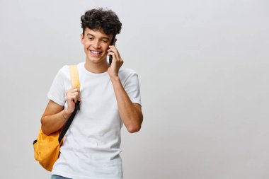 Kıvırcık saçlı canlı bir genç adam sarı bir sırt çantası taşırken telefonda konuşuyordu. Sade, beyaz bir tişört giyiyordu. Yumuşak, gri bir arka plana karşı günlük ve dostane bir hava yayıyordu.