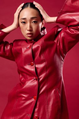 Kırmızı deri ceketli, elleri kafasında ve saçları arkada kırmızı arka planda poz veren Asyalı bir kadın. şık esmer model..