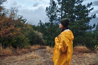 A woman in a yellow raincoat standing on a dirt path admiring the beauty of the sky during her outdoor travel adventure clipart