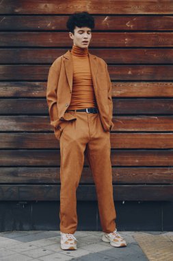 Kahverengi takım elbiseli ve balıkçı yakalı bir erkek model kırsal bir ahşap duvarın önünde zahmetsizce poz veriyor.