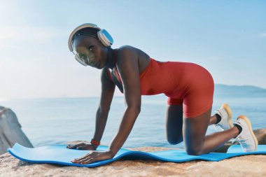 Kırmızı kıyafetli genç atletik kadın deniz kenarındaki kulaklıklarla mavi bir minderde yoga yapıyor.