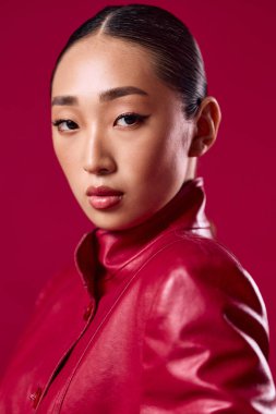 Kırmızı deri ceketli Asyalı kadın pembe arka planda at kuyruğu saçlı poz veriyor..