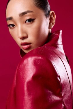 Kırmızı deri ceketli Asyalı kadın kırmızı arka planda poz veriyor gözleri kapalı ve saçları at kuyruğu şeklinde..