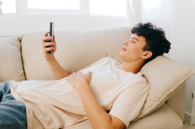 Genç bir adam bej bir kanepede uzanıyor, akıllı telefonuyla meşgul, gevşeme ve rahatlığı somutlaştırıyor. Yumuşak ve sıcak tonlarda, dijital temaslar için mükemmel, huzurlu bir ev ortamı yaratıyor.