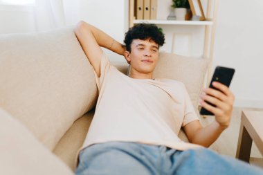 Genç bir adam, rahat bir şekilde yumuşak bir kanepeye uzanmış, rahat bir ortama dalmış akıllı telefonunu kullanıyor, oda doğal ışıkla dolu, sakin bir ortam yaratıyor.