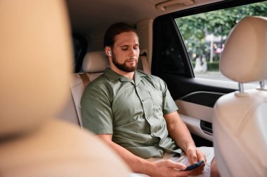 Bir adam rahat bir şekilde arabanın arka koltuğunda oturuyor, akıllı telefonuna odaklanmış, kulaklık takıyor. İçinin yumuşak, nötr bir renk paleti var.
