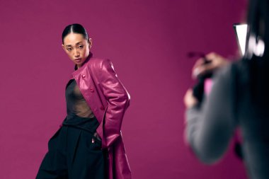 Mor deri ceketli ve pantolonlu Asyalı bir kadın, mor arkaplan önünde bir fotoğraf için poz veriyor, arkasında bir fotoğrafçı var. Moda çekimi.