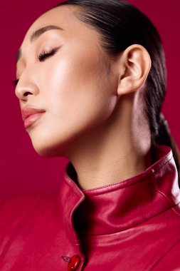 Kırmızı deri ceketli modaya uygun Asyalı esmer, gözleri kapalı, saçları at kuyruklu, kırmızı arka planda..